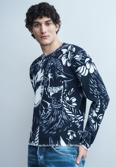 Jeune homme aux cheveux bouclés portant un pull bleu marine orné de motifs floraux blancs et de figures, associé à un jean bleu, debout contre un mur gris.