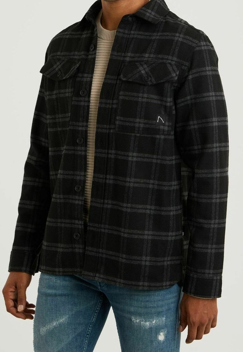Black and gray plaid shirt jacket with button closure, two chest pockets, and a subtle logo. Fabric has a textured appearance. Worn over a beige top.