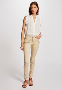 Witte mouwloze blouse met gerimpelde schouders, knoopsluiting aan de voorkant, gecombineerd met beige slim-fit jeans en bruine sandalen met een bloemaccent.