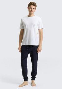 T-shirt blanc à manches courtes associé à un pantalon de survêtement bleu marine foncé avec une taille élastique et des poignets aux chevilles. Un modèle pieds nus se tient devant un fond gris.
