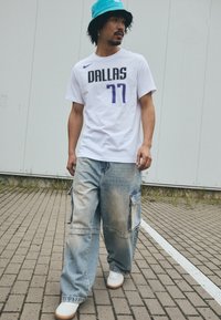 Hvid t-shirt med "DALLAS" og "77" i sort og lilla, parret med slidte blå cargo-bukser og hvide sneakers. Turkis bøttehue.