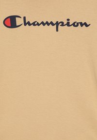Beżowa bawełniana koszulka z ciemnoniebieskim logo "Champion". Materiał ma gładką strukturę i klasyczny design z okrągłym dekoltem.