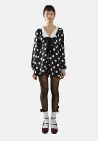 Robe noire à pois blancs, décolleté en V, col en dentelle blanche et nœud noir ; associée à des collants noirs, des chaussettes blanches et des chaussures bourgogne.