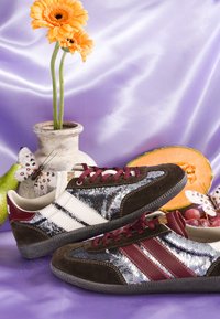 Paire de baskets à sequins avec des rayures bordeaux et blanches, fleurs orange dans un vase, papillons, poire, melon et raisins sur un tissu violet.