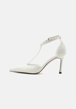 Witte pumps met spitse neus en dunne enkelriem met gesp, gladde afwerking en slanke stiletto hak op een effen achtergrond.