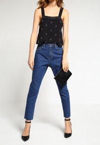 Top senza maniche nero con cuciture decorative, jeans blu a vita alta, pochette nera e tacchi neri con cinturino alla caviglia.