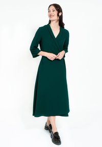 Zelené obtažené šaty s troma štvrtinovými rukávmi, výstrihom do V a splývavým strihom. V páre s čiernymi topánkami, na bielom pozadí.