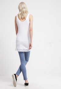 Biała koszulka na ramiączkach z koronkowym wykończeniem na hemie, w połączeniu z jasnoniebieskimi dopasowanymi dżinsami i czarnymi butami wsuwanymi. Materiał wydaje się miękki i gładki.