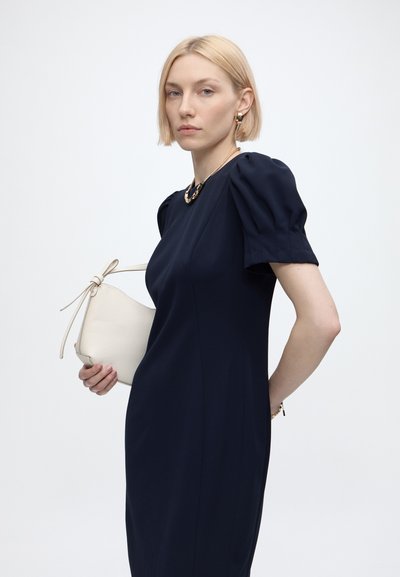 Femme blonde vêtue d'une robe bleu marine à manches bouffantes, tenant un sac à main blanc et accessoirisée avec des bijoux en or sur un fond uni.