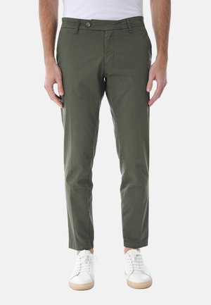 Uomo che indossa pantaloni aderenti color verde oliva e scarpe da ginnastica bianche, in piedi davanti a uno sfondo chiaro e uniforme.