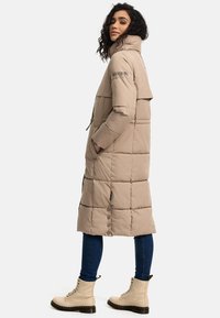 Beige padded long coat with high collar, quilted pattern, side pockets, and button accents. Paired with light-colored lace-up boots and jeans.