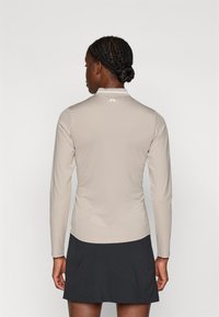 Maglia sportiva beige a maniche lunghe con finitura testurizzata e colletto bianco, abbinata a una gonna scura. Il design presenta un logo sul retro.