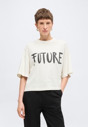 Persona con el pelo corto que lleva una camiseta blanca de gran tamaño con el texto "FUTURE" y pantalones negros, de pie con una mano en el bolsillo.