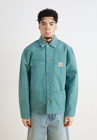 Veste de travail turquoise en coton avec un col, des boutons dorés et deux poches avant. Portée par-dessus un pantalon en denim bleu clair.