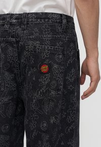 Μαύρο τζιν από denim με λευκά σχέδια ζωγραφισμένα, μεταξύ αυτών λουλούδια και αστέρια, με ένα κόκκινο και κίτρινο patch λογότυπο Santa Cruz στην πίσω τσέπη.