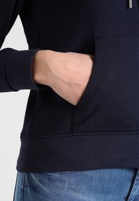 Felpa con cappuccio blu navy, dotata di tasca frontale e polsini a costine, realizzata in tessuto liscio. Presenta una zip al collo e una forma leggermente aderente.