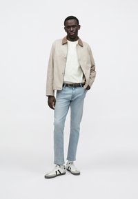 Giacca beige chiaro con colletto marrone, camicia bianca, jeans azzurri e sneakers grigie con strisce nere. Modello in piedi con una mano in tasca.