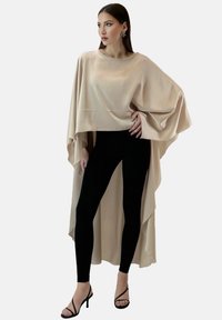 Beżowa satynowa bluzka z peleryną, z okrągłym dekoltem, zwisającymi bokami, połączona z czarnymi legginsami i sandałami na paski. Gładka faktura, minimalistyczny design.