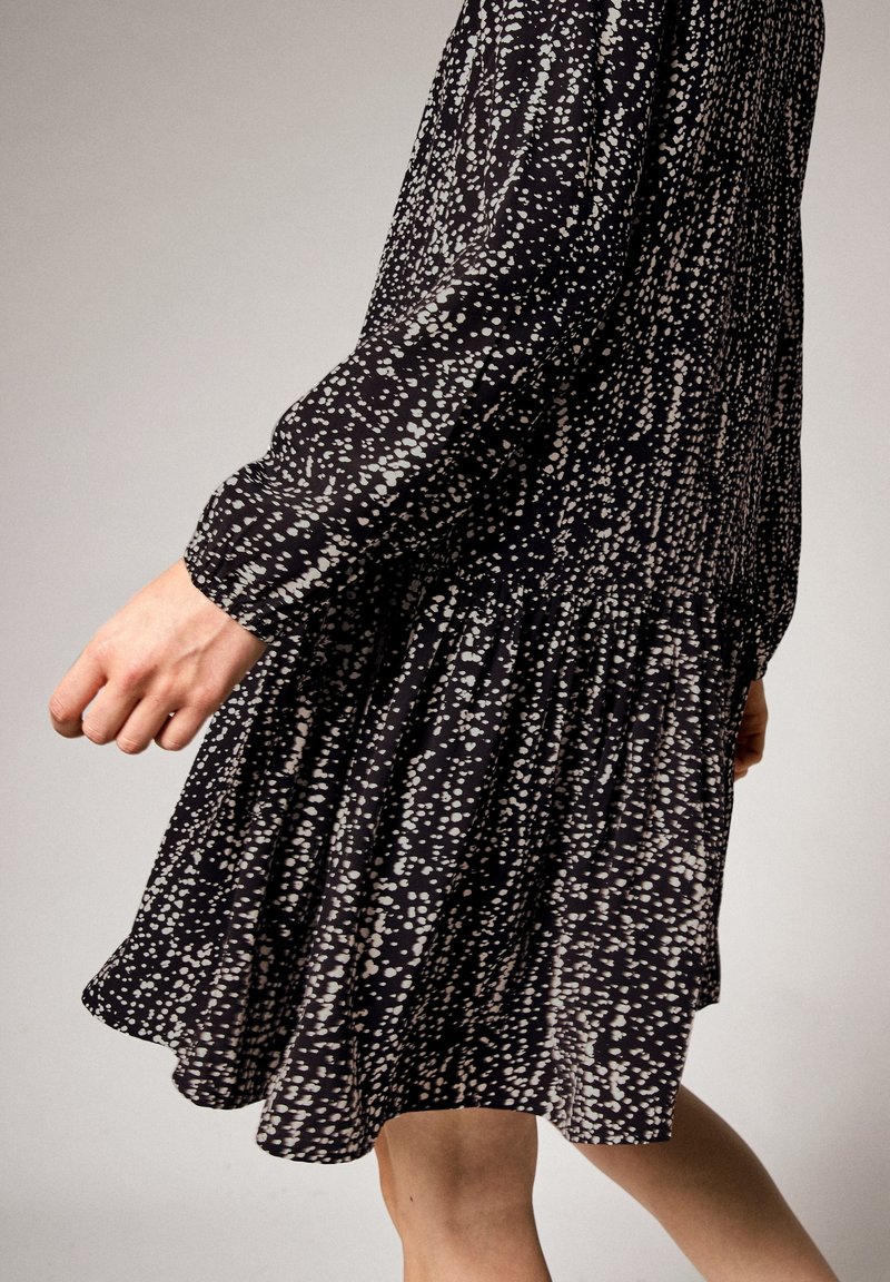 Schwarzes Kleid mit weißen Sprenkeln, langen Ärmeln, gerafftem Taillenbereich und schwingendem Saum. Leichter Stoff mit einer weichen Textur.