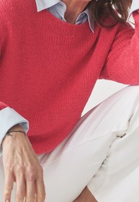 Korallenfarbener Strickpullover mit lockerer Passform und texturiertem Muster, kombiniert mit einem hellblauen, kragenlosen Hemd, das über weißen Hosen getragen wird.