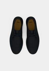 Μαύρα δερμάτινα παπούτσια δετά με καφέ εσωτερικές σόλες με τη λέξη "BOSS", τοποθετημένα δίπλα το ένα στο άλλο σε λευκό φόντο.