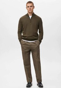 Sudadera verde oliva con cremallera, camisa blanca por dentro, pantalones beige y zapatos negros. La sudadera tiene un acabado texturizado, los pantalones son lisos.