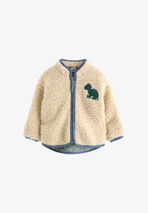 Kermanvärinen vauvan fleecetakki, jossa siniset reunukset ja vihreä dinosaurusmerkki vasemmassa rinnassa, edessä vetoketjukiinnitys.