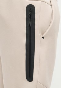 Μπεζ παντελόνι από ύφασμα με κάθετη μαύρη αδιάβροχη τσέπη με φερμουάρ στο μηρό κοντά σε μια κανονική πλευρική τσέπη.