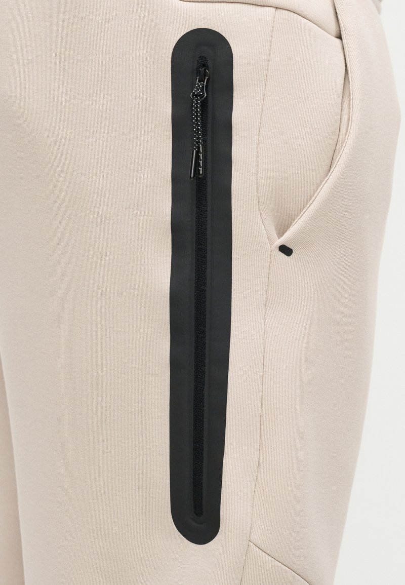 Pantalones de tela beige con un bolsillo vertical con cremallera impermeable negra en el muslo, cerca de un bolsillo lateral normal.