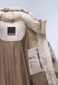 Pufferjacka med ett beige ytterlager och ett ljust rutigt innerfoder. Har en zippered ficka och varumärkestags som indikerar att materialet är fritt från dun.
