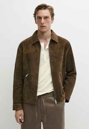 Homme portant une veste en daim marron par-dessus une chemise henley blanche et un pantalon marron ample, debout devant un fond clair uni.