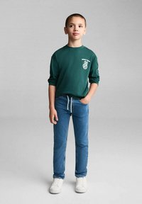 Zelené tričko s dlouhým rukávem a bílým grafickým nápisem "Skvělá nálada" a symbolem míru, spárované s modrými džíny a bílými teniskami, stojící na šedém pozadí.