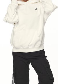 Vit bomullshoodie med en känguruficka, ribbat nederkant och svart palmträd-logga på vänster bröst. Kombination med svarta cargobyxor.