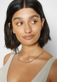Gold-tone metal necklace with a wavy design, resting on the collarbone. Model wears a light grey sleeveless top. Smooth, polished surface.