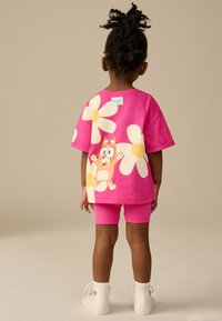 Maglietta di cotone rosa con grandi stampe di fiori bianchi e un personaggio dei cartoni animati sul retro, abbinata a pantaloncini rosa coordinati, indossata con scarpe bianche.