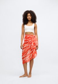 Κόκκινο διαφανές μαντήλι με λευκό χειρόγραφο κείμενο, δεμένο στη μέση, με ρέουσα σχεδίαση και ασύμμετρο στρίφωμα. Φοριέται πάνω από λευκό cropped top.