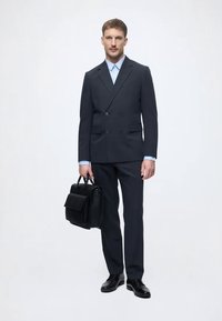 Dunkelblaues, doppelt geknöpftes Anzug mit breiten Revers, kombiniert mit einem hellblauen Hemd. Eine elegante schwarze Lederaktentasche rundet das Outfit ab.