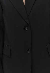 Mustat bleiserit, joissa on leikkatut kaulukset, kaksi etutaskua ja kaksi mustaa nappia. Valmistettu sileästä, rakenteellisesta kankaasta, joka on huolella muotoiltu.