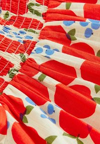 Tissu rouge et blanc avec de grands motifs de fruits rouges agrémentés d'accents bleus et verts. La texture montre des sections plissées et froncées.