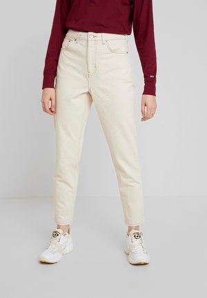 Persoon draagt high-waist off-white jeans, een bordeauxrode lange mouwen shirt en witte sneakers, staand tegen een effen grijze achtergrond.