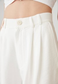 Pantalones blancos de cintura alta con trabillas y cierre de botón, llevados por una persona con el abdomen visible sobre un fondo liso.