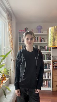 Svart hettejakke med glidelås over en svart skjorte, sammen med svarte bukser. Innstillingen har en bokhylle og en stor gyllen lampe.