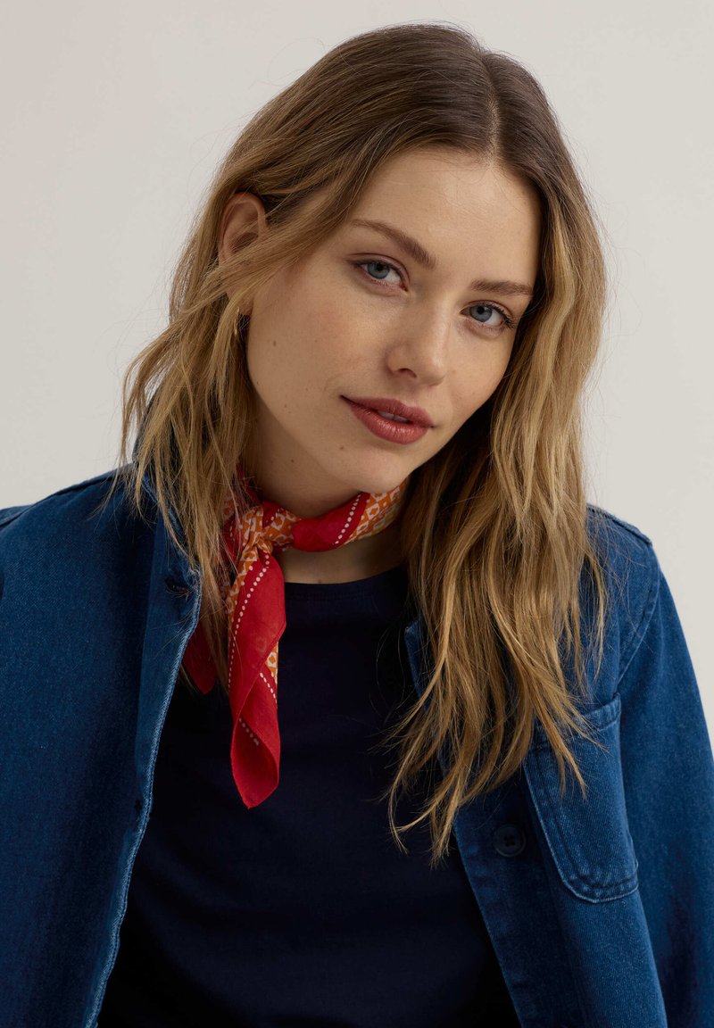 Jeune femme aux longs cheveux blonds portant un foulard rouge à motifs autour du cou, une chemise bleu marine et une veste en denim bleu sur un fond uni.