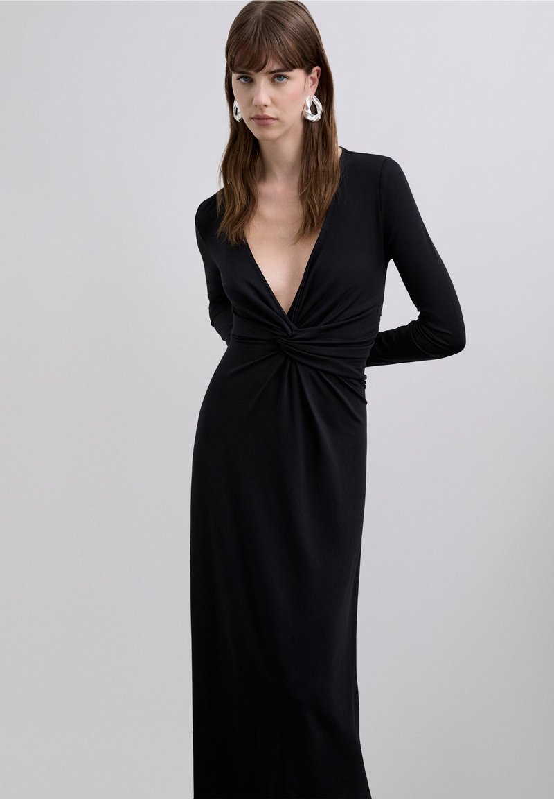 Robe noire à manches longues avec un décolleté plongeant en V, un détail torsadé sur le devant et un tissu lisse, accompagnée de grandes boucles d'oreilles argentées.