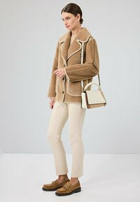 Bruine pluche jas met witte afwerking, crèmekleurige top, op maat gemaakte crèmekleurige broek en bruine suède loafers met gesp, aangevuld met een kleine tas.