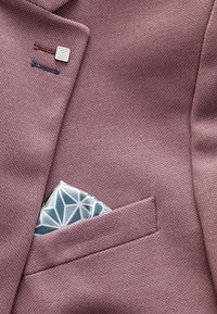 Bordový texturovaný blazer s jednou kapsičkou, která má geometrický modro-bílý vzor. Stříbrný knoflík nad kapsou.