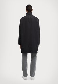 Černý, oversized kabát s vysokým límcem, dlouhými rukávy a hladkým povrchem, doplněný o světle šedé džíny a bílé boty.