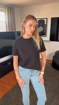 Tânără cu păr blond lung, purtând un tricou negru și blugi albastre deschis, stând într-un sufragerie cu televizor și opere de artă înrămate.