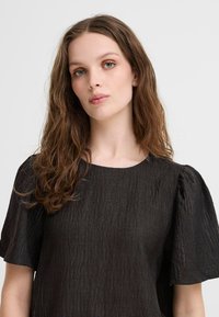 Blusa nera con texture, dotata di maniche corte a sbuffo e scollatura rotonda, realizzata in un tessuto leggero con un leggero brillantezza.