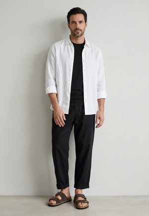 Muž stojící u zdi, nosící bílou košili s knoflíky přes černé tričko, černé kalhoty a hnědé sandály s otevřenou špičkou a s přezkami.